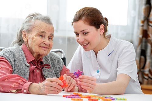 Young CNA in white scrubs helping elderly woman with color puzzle