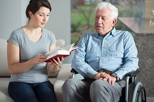 Young woman sitting next to elderly man in wheelchair reading him a book